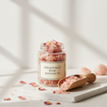 Himalayan pink bath salts in glass jar with rose petals on white background